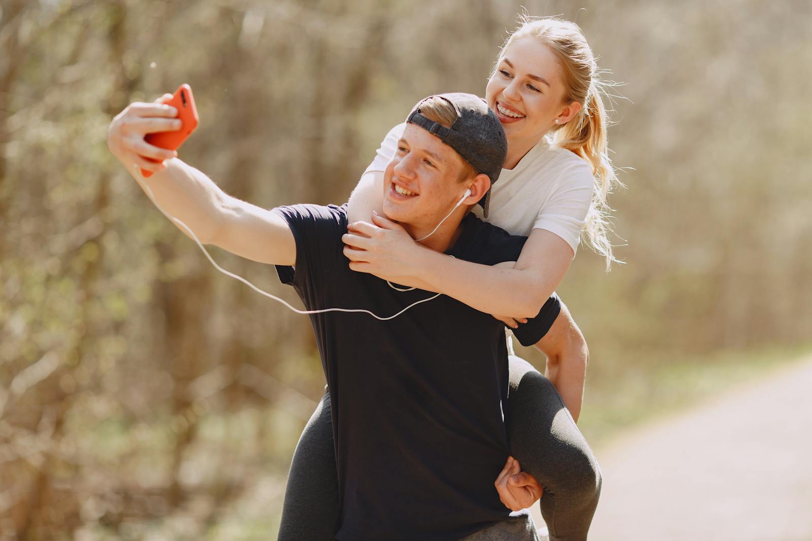 A joyful couple taking a selfie while enjoying a piggyback ride in a sunny park.